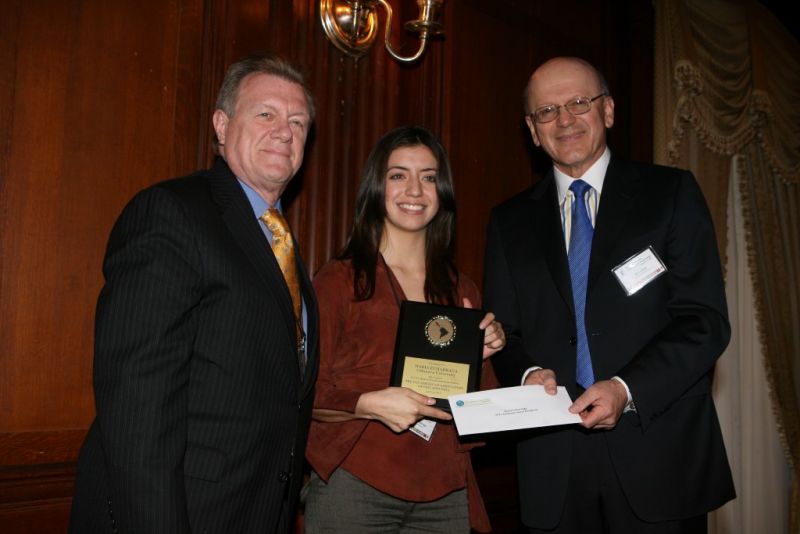 Two men in suits taking a photo with a woman holding an award, while one of the men hands her a letter.